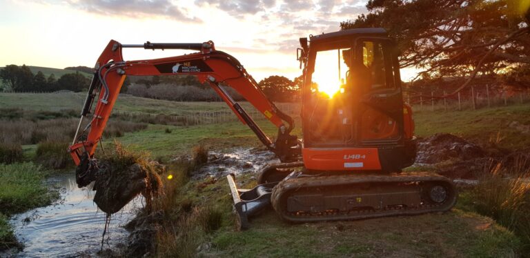 Picture of the excavator with Janus in it before the sunset in the background, sun shining through the cabin.