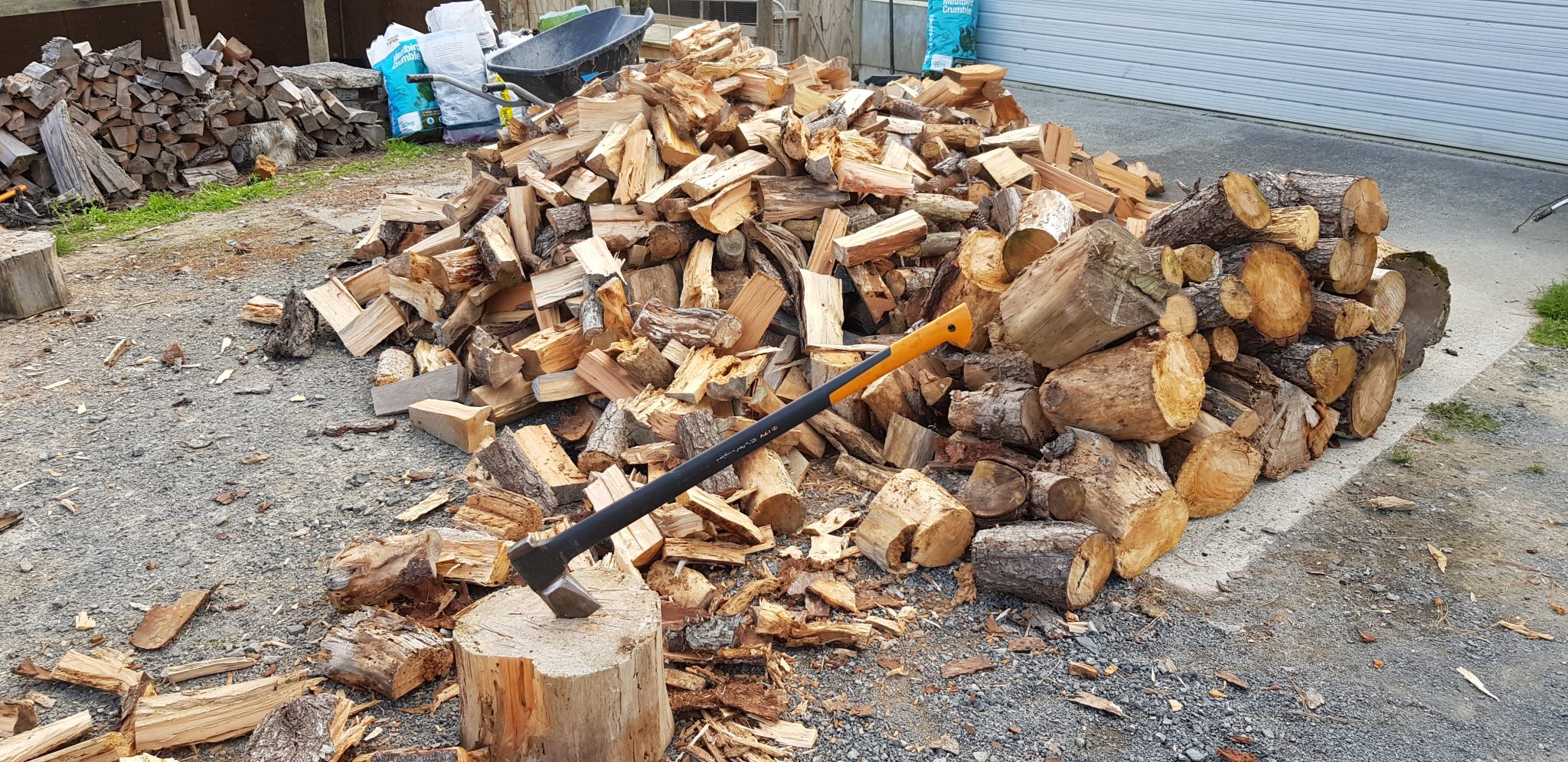 Piled up split firewood and some big chunks of wood in front of it with a chopping block