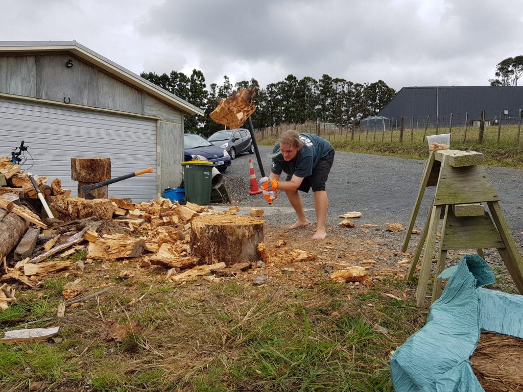 Man splitting firewood with splithammer, barefeet, surrounded by pieces of wood
