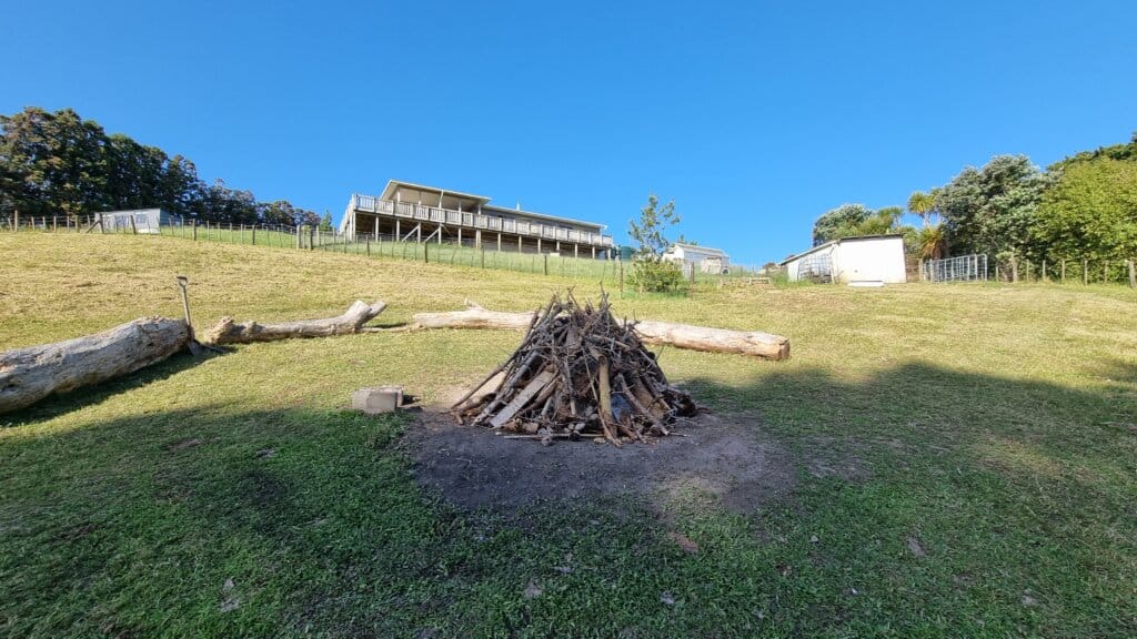firewood pile stacked up in the paddock with hill sloping up with house higher up in the background, blue sky
