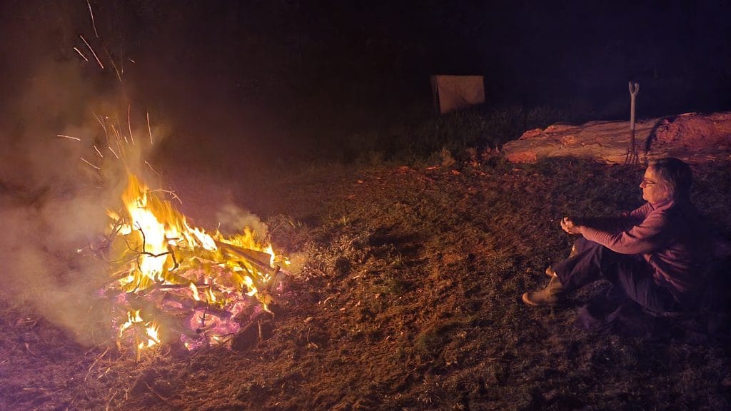 A bonfire in a dark paddock at night with a person sitting in some safe distance to the fire