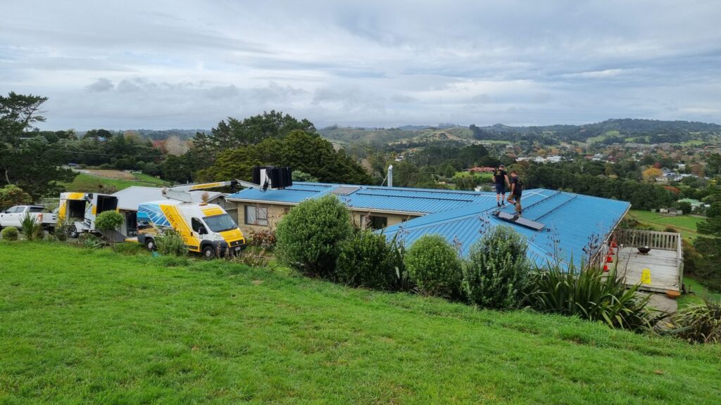 Yellow van with boom has lifted solar PV panels onto the roof. Rails have been installed across the roof and two people are busy installing black, flat solar panels.