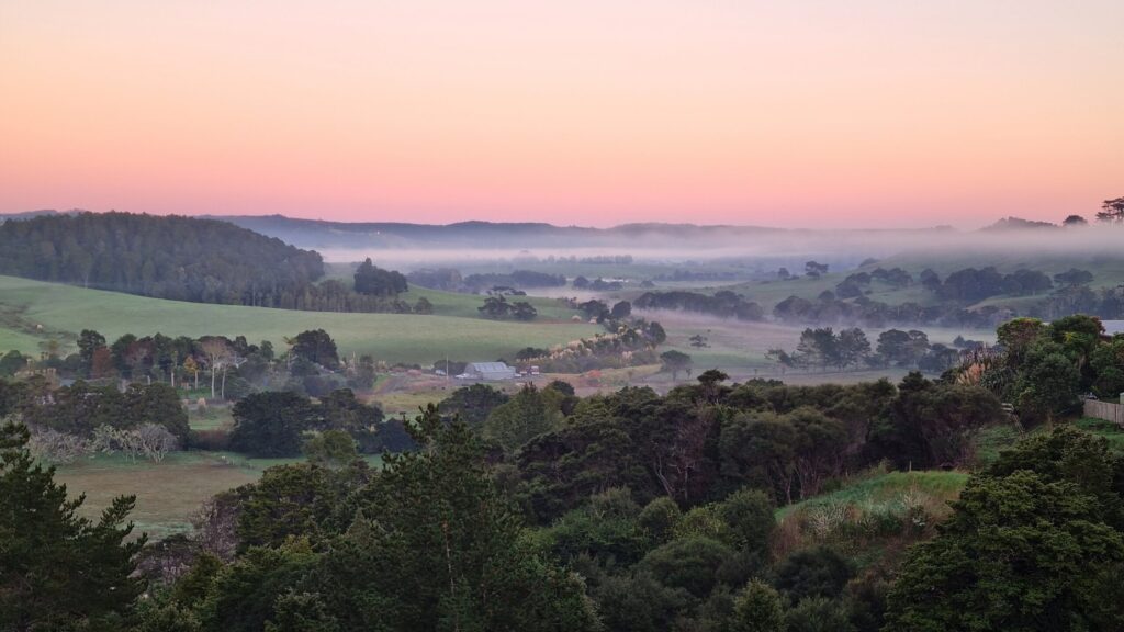 A valley with fields and rows of trees with lingering fog in between, orange, blue-grey mist and light in the sky in the background